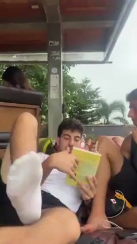 Two young men are relaxing on leather beanbag chairs on a covered balcony. The man on the left is wearing a white t-shirt and black shorts with a white sock on his left foot. He's holding a yellow book and pointing at a page, seemingly reading aloud to the other man. The man on the right is wearing a black tank top and black shorts. He has his right arm resting on his knee and his left hand on his chin, listening. A remote control sits near his leg on the beanbag. In the background is a woman with dark hair sitting in a chair and a lush green landscape under a cloudy sky. The balcony has a wooden roof and a metal railing.