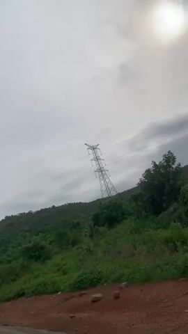 The video shows a car moving along a dirt road next to a steep, tree-covered hill. A tall electrical pylon is visible on the hillside, with power lines stretching across the frame. The sky is overcast with the sun faintly visible through the clouds. The road is red dirt and appears to be in a rural or natural setting.