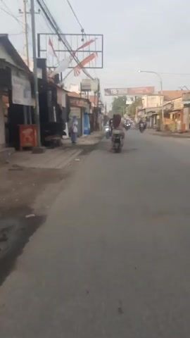 The video captures a street scene with several people on motorcycles. On the left side of the road, there are shops and buildings. A person in a reflective vest stands near the side of the road. The road is paved, and there are overhead wires and banners. The overall atmosphere appears to be that of a busy street in a populated area.