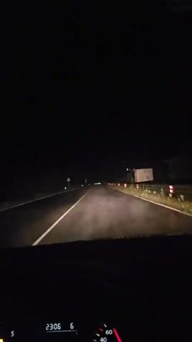A first-person view from inside a car driving on a dark, wet road at night. The car's headlights illuminate a portion of the road ahead, showing some mist or fog just above the road surface. Road signs are visible on the left side, and a guardrail with red markers is on the right. The dashboard is visible at the bottom of the frame, displaying the time as "23:06" and a speed indicator. The car appears to be driving through a rural or unlit area.
