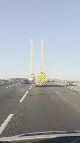 The video shows a perspective from inside a vehicle driving on a highway over a large cable-stayed bridge. The sky is clear and blue. In front, there are several vehicles on the road, including a black car, a white truck, and a yellow and red striped van that appears to be some kind of emergency or service vehicle. The bridge has two tall, white pylons supporting numerous cables that fan out to hold up the road deck. The roadway is asphalt with white lane markings. On either side of the road are railings and barriers. Beyond the bridge, a landscape with some buildings and vegetation is visible. The camera is moving forward, showing the vehicles ahead and the immense structure of the bridge.