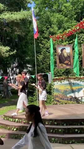 The video shows an outdoor event likely taking place in Obninsk, Russia, on a sunny day. A group of young people are on a raised wooden stage decorated with greenery and flags. A Russian flag flies prominently behind them. To the right of the stage, there are two green flags and a framed portrait of what appears to be an older man with a beard, possibly a historical or cultural figure. A painted backdrop depicting a landscape scene with a town and a river is visible behind the performers.

On the stage, a young woman in a white t-shirt and white shorts with pink sneakers is the central performer. She is gesturing with her hands as if speaking or singing. To her left, a group of three other young people, two women and a man, are also on the stage, appearing to be participants or performers. The woman on the far left wears a black t-shirt and a white patterned skirt, while the woman next to her wears a white dress. The man is in a black t-shirt and light blue jeans. Their body language suggests they are engaged in a performance or activity.

In the foreground, two individuals are partially visible. One is a woman wearing a white long-sleeved shirt and a green cap, walking past the stage. Another person in a red dress is also visible in the foreground, walking away from the camera.

The overall atmosphere appears festive and celebratory, possibly a cultural event or a school performance given the young age of the participants and the decorative elements. The sunlight suggests it is daytime. There is no visible text in the video.