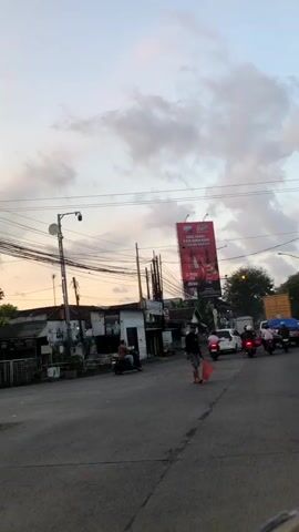 The video shows a street scene with several vehicles and people. The time of day appears to be dusk or dawn due to the lighting. There are motorcycles and cars on the road. A person is walking across the street, carrying a red object. In the background, there is a large billboard with red lettering and images. There are also buildings and a lot of overhead wires, possibly for electricity or telecommunications. The sky is cloudy.