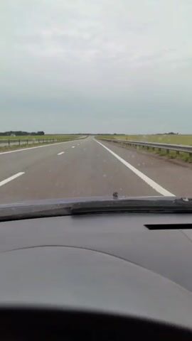 The video shows a first-person view from inside a vehicle, looking out through the windshield at a highway. The highway has two lanes in the visible direction, separated by a white line, and a solid white line on the right edge. Guardrails line both sides of the road. The sky is overcast and gray, and the landscape on either side of the highway appears to be flat farmland or fields. The vehicle's dashboard is visible in the foreground. The car is in motion, indicated by the steady forward perspective of the road. No other vehicles or people are visible.