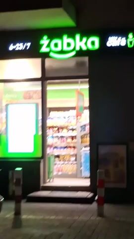 The video captures the exterior of a Żabka convenience store at night. The store has a bright green neon sign that reads "żabka cafe" with the hours "6-23 / 7" visible to the left. The entrance is a glass door, and through it, shelves stocked with various products are visible. In front of the store, there are two red and white striped bollards on the sidewalk. To the right of the entrance, there's a poster displayed outside, and another window offers a glimpse of the store's interior. The street pavement is visible in the foreground.
