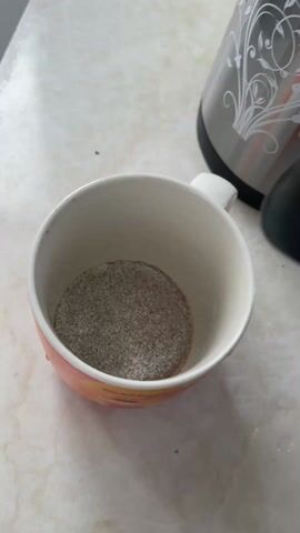 A cup on a marble surface contains what appears to be instant coffee powder. Hot water is poured into the cup from a black electric kettle. A stainless steel thermos sits in the background.
