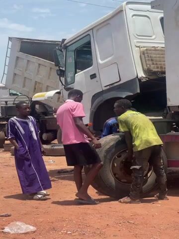 Three men are working on a large truck tire on a red dirt surface. The truck's cab is tilted forward. One man wears a long purple robe, another a pink shirt and black shorts, and the third a yellow shirt and dark pants. They are all barefoot or wearing sandals. The truck is white, and another truck is visible in the background. The sky is blue with scattered clouds.