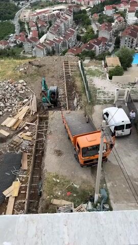 The video shows an aerial view of a construction site. An excavator is digging a trench. A flatbed truck is parked nearby, and a white van is also present. Two men are standing near the van. There are piles of debris and construction materials on the ground. In the background, there are residential buildings and a swimming pool. The overall scene suggests ongoing construction work in a suburban or urban environment.