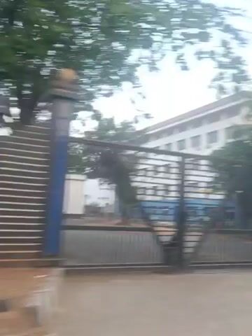 The video shows a blurry street view with buildings and trees. A black fence with blue accents is visible in the foreground. The fence has vertical bars and horizontal slats. A large, modern building with glass windows and the letters "TSP" on it is visible behind the fence. Another building, which appears to be a modern office building, is also seen. The sky is overcast. There are some power lines visible. Some people on motorcycles are seen passing by. The video is shaky, suggesting it was filmed from a moving vehicle.
