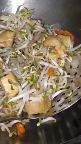 The video shows a close-up, overhead shot of a metal wok filled with bean sprouts, diced tomatoes, and cubed tofu in a dark, savory broth. A dark slotted spoon is seen stirring the ingredients. The food appears to be in the process of being cooked or stirred. The lighting is overhead and casts some glare on the metal wok.