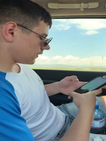 The video shows two young men in a car with a sunroof. At the start of the video, one of the men is in the passenger seat, looking at his phone. The man driving has a shaved head, a beard and wears a black t-shirt. He’s driving on a sunny day on a paved road through what seems to be countryside. An air freshener hangs from the rearview mirror. Loud music can be heard.