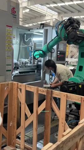 A person stands in front of an industrial lathe, making notes on a clipboard.  The lathe is a large metal machine with multiple tools and a rotating chuck. An articulated robot arm, painted green, extends from the right side of the frame toward the lathe. A wooden crate is in the foreground, partially obstructing the view. The industrial space appears well-lit.