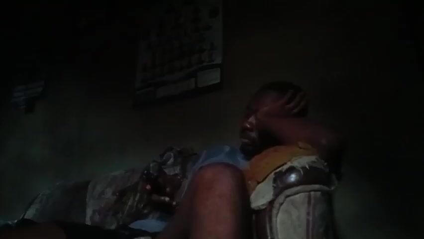 A man is sitting on a couch with his hand on his forehead. He is holding a phone. The room is dark, with a picture on the wall behind him. There is a window with bars on it.