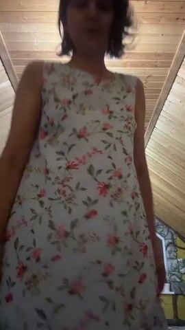 A woman stands in front of a camera, wearing a sleeveless, light-blue floral dress. The dress has a ruffled hem. She has short, dark hair and appears to be indoors, possibly in a room with a sloped wooden ceiling. There is a bright light directly above her. She moves her hips and places her hands on her hips.