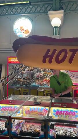 A man in a green shirt is standing near a claw machine filled with stuffed animals. In the foreground, there are several arcade games with coins visible in their trays. In the background, people are walking and playing games. The scene appears to be an indoor arcade or amusement park.