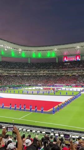A soccer stadium is shown at dusk with green lights illuminating the stadium roof. A large flag, red, white, and blue, is spread across the field. People in blue uniforms stand on the edge of the flag, creating a border. Many spectators are in the stands, and some in the foreground are holding up phones to record the event.