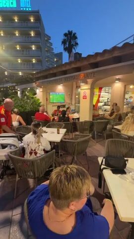 The video shows an outdoor seating area of a restaurant with several patrons. The time of day appears to be dusk, as indicated by the twilight sky and the lights on in the restaurant. In the background, a television screen is visible, displaying a soccer match. People are seated at tables, some eating and drinking, while others appear to be watching the game. The restaurant has an awning with the text "CAFETERIA RESTAURANTE" and "LA COLMENA". Spanish flags are displayed on the building. In the foreground, a person wearing a blue shirt is seated facing away from the camera. The atmosphere seems relaxed and social, with people gathered to enjoy a meal and a sporting event.