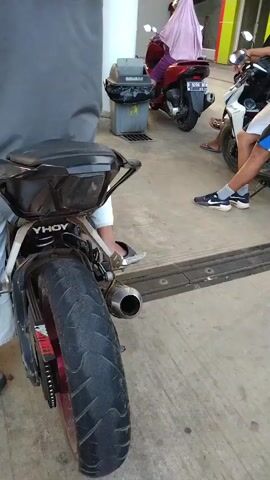 The video shows a close-up of a motorcycle's rear tire, with the camera then panning to show other motorcycles and people at what appears to be a gas station. Several people are seated on or standing near their motorcycles. In the background, there are gas pumps and a building. The ground is a concrete surface. The lighting suggests it is daytime, possibly late afternoon.