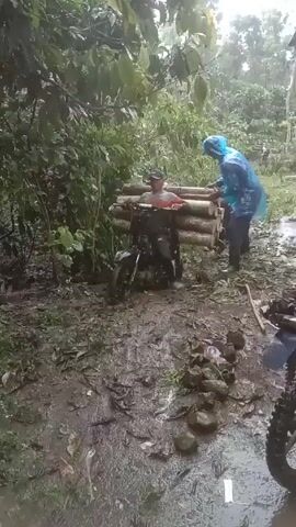 The video shows two people, one riding a motorbike in a muddy area surrounded by vegetation. The first person is seated on the motorbike, facing the camera, with a stack of logs secured to the front of the bike, appearing to brace against them. The second person, wearing a blue poncho, stands beside the motorcycle. The ground is covered in thick mud, leaves, and sticks. Another motorbike is partly visible in the bottom right corner of the frame. The surroundings are dense with green trees and foliage, suggesting a wooded area or rainforest environment, and it appears to be raining or recently rained.