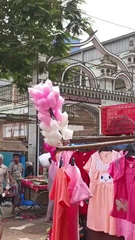 The video depicts a bustling street market scene with several vendors and pedestrians. In the initial shot, there's a cart with pink and white cotton candy, as well as a display of children's clothing. In the background, there's a mosque. The camera then pans, revealing more market stalls covered by makeshift awnings selling clothing and fabric. People are browsing, walking, and interacting with the vendors. Some individuals are wearing masks. The overall environment is crowded and appears to be a typical outdoor marketplace.