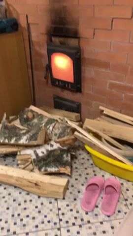 A static shot reveals a brick fireplace in a room. The fireplace door is closed, but the interior is visible, glowing with orange light, presumably from a fire burning inside. Above the fireplace, there's a noticeable soot mark on the brick wall. In front of the fireplace, there's a pile of chopped wood, including some birch logs with their characteristic white bark. To the right of the wood, a yellow plastic tub contains more pieces of wood, likely kindling. On the tiled floor in the foreground, a pair of pink slippers sits neatly. A wooden cabinet is visible to the left of the fireplace. The overall setting suggests a rustic or cozy interior, possibly a country house or cabin.