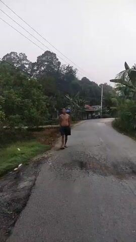 The video shows a man walking on a road. He is shirtless and wearing shorts. The road is paved and appears to be in disrepair, with several potholes visible. The man is walking towards the camera. There is vegetation on either side of the road, and trees can be seen in the background. The sky is overcast.