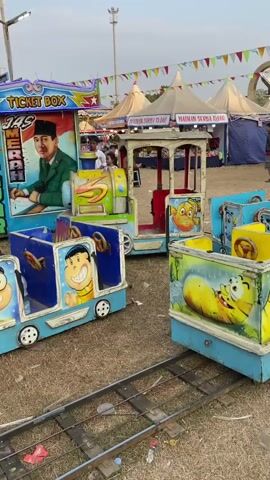 The video shows a small amusement park ride, likely a kiddie train, in an outdoor setting. The train cars are brightly colored and decorated with cartoon characters, including what appears to be Doraemon. The ride is set up on a grassy area with train tracks visible in the foreground. Behind the ride, there are various stalls or booths, some with tents and banners, suggesting a fair or festival. People can be seen in the background, and there is a ticket booth with a picture of a man in a suit. The overall atmosphere appears to be casual and festive.