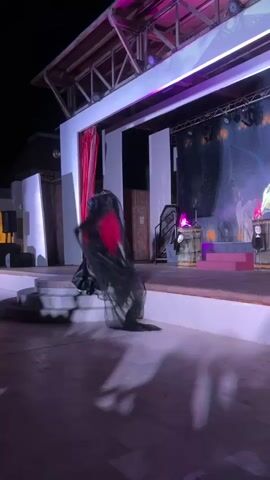 A person in a black veil and red accents, possibly a costume, is dancing or performing on a stage at night. The stage has theatrical lighting and a backdrop depicting a scene with what appears to be a castle and figures. There are props such as barrels, a coffin-like structure, and possibly statues of angels or mythical beings. Smoke or fog effects are visible on stage. The ground in front of the stage is paved, and shadows of figures are cast upon it. The overall atmosphere suggests a dramatic or theatrical performance.