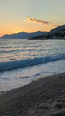 A tranquil beach scene unfolds as gentle waves lap against a pebble-strewn shore. The setting sun casts a warm, orange glow across the horizon, illuminating the distant, silhouetted mountains. The calm sea reflects the soft hues of the sky, creating a peaceful and serene atmosphere. Small boats are visible in the distance, suggesting a coastal town or harbor. The overall mood is one of serenity and natural beauty.