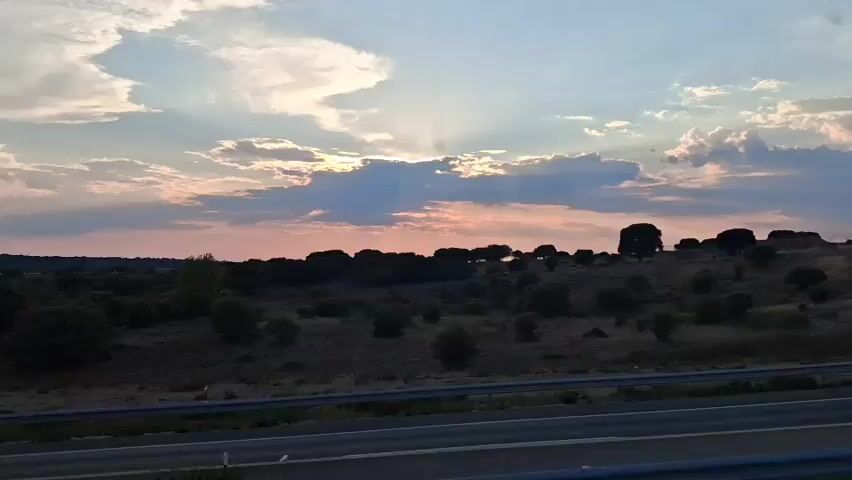 A handheld video shows a silhouetted landscape under a sky with scattered clouds and sunbeams. The foreground features a dark road or highway with a white line, suggesting movement. The midground consists of a gently sloping, darkened terrain dotted with various trees and shrubs. The background is dominated by a sky with pinkish and bluish hues, indicative of either sunrise or sunset, with clouds partially obscuring the sun. A single black car is briefly visible driving on the road in the first second of the video. The overall scene is a natural landscape viewed from a moving vehicle.