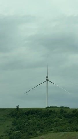 A single white wind turbine stands on a grassy hill under a cloudy sky. The hill is covered in green bushes and a small tree is visible on the left side of the frame. The wind turbine's blades are turning. Electronic music plays in the background.