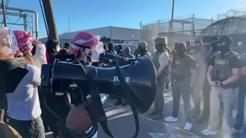 In a daylight outdoor scene, a group of protesters faces a line of law enforcement or security personnel. The setting appears to be outside an industrial or correctional facility, characterized by tall fences topped with barbed wire in the background. The sky is clear blue.

On the left, several protesters are visible. One individual, wearing a white shirt, a red and white keffiyeh, and clear safety goggles over a black face mask, holds a black megaphone to their mouth and appears to be leading chants. Another protester next to them also holds a megaphone. Both megaphones bear the brand name "PYLE" and "PyleUSA.com". The protesters are actively chanting, with visible text indicating "F*ck ICE!" and Spanish phrases like "Jungle la negra" and "Escuela de Perqueria".

Opposite the protesters, a line of approximately ten law enforcement or security personnel stands in formation, observing. Most are wearing dark tactical gear, including helmets, balaclavas covering their faces, and ballistic vests. Some wear khaki-colored trousers, while others are in darker pants. Several personnel have "ERO" emblazoned on their vests, likely referring to ICE Enforcement and Removal Operations. One officer in the center has a shield or vest with "POLICE" visible on it. Their demeanor appears stoic and prepared, providing a strong contrast to the vocal protesters. A utility pole is visible in the foreground on the left.
