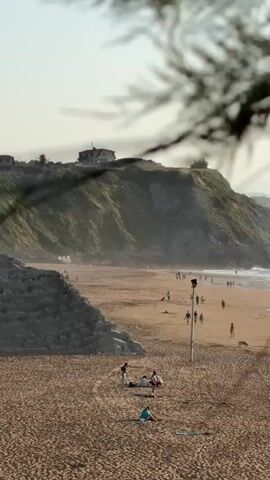 The video shows a view of a beach with several people walking and sitting on the sand. In the background, there is a hill with a house on top, and the ocean. The lighting suggests it is daytime, possibly late afternoon or early evening. The camera appears to be stationary, looking down at the beach from an elevated position. Some foliage is visible in the foreground, partially obscuring the view.