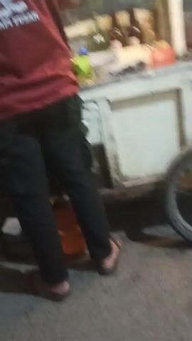 This video shows a person wearing a red t-shirt with a cartoon character and text on the back. The person is standing next to a food cart. The cart appears to be a mobile food vendor, possibly selling noodles, as indicated by the text on the cart. The cart is stocked with various items, including bottles, containers, and possibly food ingredients. In the background, there is a dimly lit street with a bamboo pole and a house decorated with lights. The overall scene suggests a nighttime setting, likely in an urban or semi-urban area where street food is being sold.