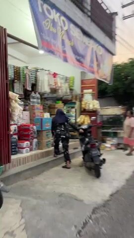 The video shows a person in a black hijab and patterned clothing entering a store. The store appears to be a small convenience store or warung. There are shelves stocked with various items, including bottled water, packaged goods, and what appear to be disposable cups. A motorcycle is parked in front of the store. Another person wearing a pink shirt and dark pants is seen walking away from the store carrying a large water jug. The lighting suggests it is either evening or a dimly lit indoor space. The overall scene depicts a typical transaction or shopping experience at a local neighborhood store.