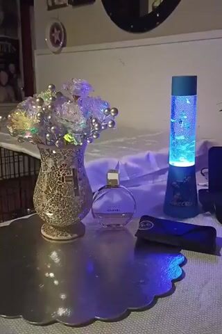 The video shows a still life arrangement on a table. The objects include a decorative vase with artificial flowers, a bottle of Chanel Chance perfume, a pair of Ray-Ban sunglasses, and a blue lava lamp. The lava lamp is illuminated and appears to contain small, fish-like objects that are moving within the liquid. The table is covered with a white cloth, and the background shows a wall with framed pictures and a mirror. The lighting is dim and appears to be coming from the lava lamp, casting blue and purple hues on the surrounding objects.