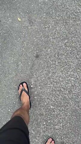 The video begins with a close-up shot of a person's feet wearing black flip-flops, walking on a grey asphalt road. A small yellow leaf is visible on the ground. The camera then pans up to a stone wall beside the road, which has blue graffiti on it, clearly displaying the word "Ljubav" (meaning "Love" in Serbian/Croatian). Finally, the view opens up to a breathtaking panorama of the Bay of Kotor in Špiljari, Montenegro. Lush green mountains descend into a deep blue bay dotted with several boats and ships. A town with numerous buildings is nestled along the coastline. The sky is partly cloudy, with patches of blue visible. The scene suggests a daytime setting with pleasant weather.