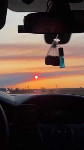 The video shows a first-person perspective from inside a car driving on a road in Vyselki, Russia, during sunset. The dashboard of the car is visible in the foreground, with a rearview mirror and two air fresheners (one appearing to contain coffee grounds, the other a blue liquid) hanging from it. The silhouette of a person's head is briefly seen on the left side of the frame.

Through the windshield, a stunning sunset dominates the view. The sky is painted in vibrant shades of orange, red, and pink, with some dark clouds stretching across the horizon. A bright, round red sun is positioned low in the sky, directly ahead.

The car appears to be on a highway or a main road, with other vehicles visible in the distance. The surrounding landscape consists of what looks like open fields or rural areas, with scattered trees and utility poles lining the road. A faint road sign can be seen on the right side of the road. The overall impression is one of peaceful travel during a beautiful evening. A voice, possibly from the car's radio or another passenger, can be heard speaking Russian, with the word "безопасно" (safely) audible.