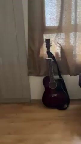 The video appears to be a short tour of an interior space, likely a home. The camera pans across a room with light-colored wooden flooring, starting from an acoustic guitar leaning against a wall near a window covered with a translucent curtain. The camera then moves to show a closed closet door, followed by another white door. A small black mat is visible on the floor near the doors. The wall next to the second door features electrical switches, outlets, and a framed photograph of three people. The camera briefly reveals a partial view of another room through a doorway, with a dark object, possibly a suitcase or vacuum cleaner, partially obscuring the view. The audio appears to include background sounds of conversation or speech in an unidentified language.