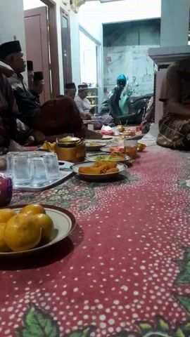This is a horizontal, medium-shot video filmed indoors, likely in a home in Panjalin Kidul, Indonesia. The scene appears to be a gathering of men, many of whom are wearing traditional Islamic attire, including peci caps. They are seated on the floor around a low table laden with food and drinks, suggesting a meal or communal event.

In the foreground, a plate of oranges sits on a red, patterned tablecloth. Next to it, a packet of "Jolly" brand tissues is visible. Further back, plates of food, glasses, and a golden teapot are arranged on the table. The men are engaged in conversation and appear relaxed. One man in the foreground is speaking animatedly, with a wide smile. The lighting suggests it is daytime, with natural light coming from an open doorway in the background. A motorcycle is parked inside the room, adding to the domestic setting. The overall mood seems convivial and social.