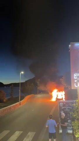 A car is on fire on a road at night. There is a lot of smoke and flames coming from the car. Two people are standing on the sidewalk, watching the fire. There is a body of water in the background, and mountains. A sign for a restaurant is visible on the right side of the screen.