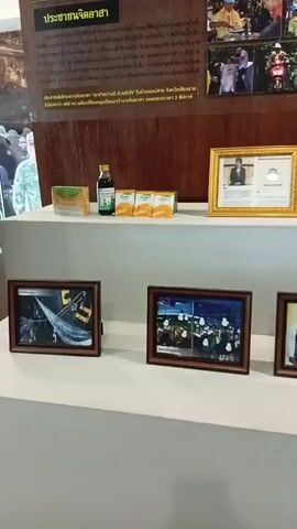 The video shows a display of items on shelves against a wooden wall. The wall features text and several framed photographs. The top shelf has several items including a small green bottle, a box, and some yellow packets. To the right of these items is a gold-framed picture of a person. The shelf below has several framed pictures, including one of a group of people, and what appears to be a rolled-up item that is white and patterned. Further to the right, there is a yellow hard hat. The environment appears to be indoors, possibly a museum or exhibition space.