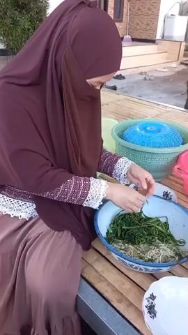 The video shows a person wearing a maroon hijab and a brown skirt, sitting on a bamboo mat. They are in front of a large white bowl filled with green leafy vegetables. The person is holding some of the vegetables and appears to be preparing them, possibly by trimming or separating them. To the right of the bowl are two plastic baskets, one blue and one pink. In the background, another person wearing a blue hijab is visible, holding a pink container. They are standing on steps outside a building. The environment suggests an outdoor setting, possibly a backyard or patio, with some vegetation visible on the left.