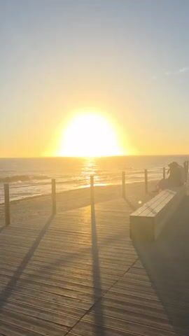 A static, vertical video captures a beach scene at sunset. The sun, a bright, diffused orb, sits low on the horizon, casting a golden glow over the ocean and a wooden boardwalk. Waves gently lap the shore. On the boardwalk, a long, wooden bench is positioned towards the right side of the frame. Two figures, a couple, are seated on the bench, facing the ocean. The person on the left is leaning their head on the shoulder of the person on the right. The scene is peaceful and romantic, with the warm light of the sunset creating a serene atmosphere. The boardwalk extends forward, its wooden planks casting long shadows in the fading light. The sky is a clear, soft blue, transitioning into a pale yellow near the horizon.