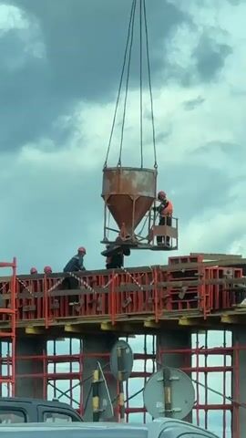A daytime scene captures several construction workers engaged in concrete pouring at a building site under an overcast sky. A large, funnel-shaped concrete bucket, suspended by chains from an unseen crane, is being lowered towards a concrete structure.

One worker, wearing a red hard hat and an orange high-visibility vest, stands on a small platform attached to the concrete bucket, appearing to operate controls. Another worker, also in a red hard hat and dark uniform, stands on the construction level, using a long-handled tool to guide the bucket's positioning. A third worker in a dark uniform and hard hat is also visible near the base of the bucket, assisting.

The construction level features extensive red/orange metal formwork and wooden planks, indicative of preparations for a concrete pour. Other workers in hard hats can be seen in the background on the same level. Partially visible road signs and the top of a vehicle are present at the bottom of the frame, suggesting the construction is occurring near a roadway. The workers appear focused on coordinating the precise placement of the concrete bucket.