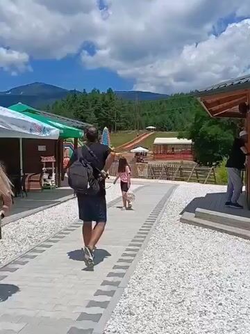 A man with a backpack is walking away from the camera down a paved path. He is wearing a dark shirt and shorts. To his left, a young girl in a pink shirt and black shorts is walking toward the camera. She is carrying a white bag. Behind her, an inflatable slide is visible. To the right of the path, there is a covered outdoor seating area with tables and chairs. In the background, there are trees and mountains. The sky is blue with white clouds.