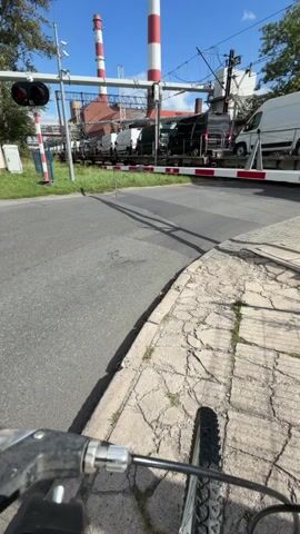 The video opens with a first-person perspective of someone on a bicycle. The camera is angled downwards, showing the handlebars and front wheel of the bicycle. In the background, a train is passing through a railway crossing. The train cars are carrying several white delivery vans and some green trucks. The railway crossing gates are down, indicating that the train has priority. There are red and white striped poles marking the crossing, and a traffic signal with red lights is visible. In the far background, two tall industrial chimneys with red and white stripes rise into a bright blue sky. The environment appears to be an urban or industrial area, with asphalt roads, a cracked concrete sidewalk, and some greenery. The sun is shining, casting shadows. The video lasts for a few seconds and shows the train continuing to move past the crossing.