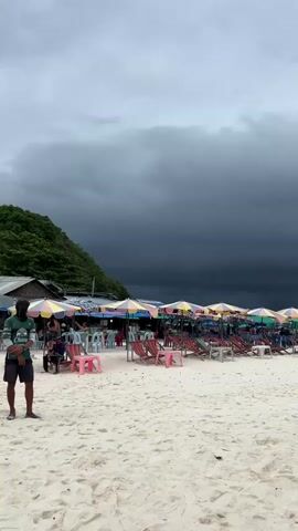 The video shows a beach with dark, ominous storm clouds gathering overhead. In the foreground, there is white sand and a few pieces of trash. A person in a green shirt and shorts stands on the left side of the frame, facing the camera. Behind them, a row of colorful umbrellas and beach chairs are set up. Several people are seated at tables and chairs under some of the umbrellas. To the left, a small building with a tin roof is visible, nestled against a lush green hillside. The beach appears to be a commercial area, with many umbrellas and chairs set up for rent. The overall mood of the video is one of impending weather, with the dark clouds creating a sense of drama.