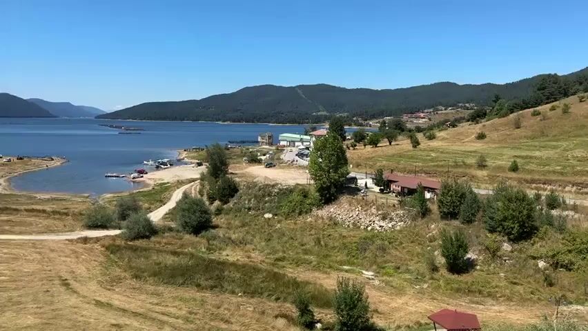 This is a landscape video with a wide shot of a lake surrounded by rolling hills and mountains. The lake is a deep blue, and there are several boats docked along the shore. The hills are covered in green grass and trees. The sky is clear and blue. In the foreground, there is a dry, grassy field with a few scattered trees. There is a dirt road winding through the field. In the middle ground, there are a few small buildings, including a red-roofed house and a larger building with a green roof. There are also several cars and RVs parked near the lake. In the background, the lake stretches out towards the horizon, with more tree-covered hills in the distance. The video appears to have been shot on a sunny day.