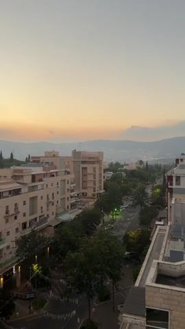 The video shows a wide shot of a city street lined with trees and buildings. The sky is a soft gradient of orange and yellow, suggesting either sunrise or sunset. In the foreground, there are apartment buildings with visible windows and balconies. A street is visible below, with trees lining one side. There are also some streetlights and decorative lights strung across the street. In the background, more buildings and hills can be seen in the distance. The overall atmosphere is peaceful and serene.
